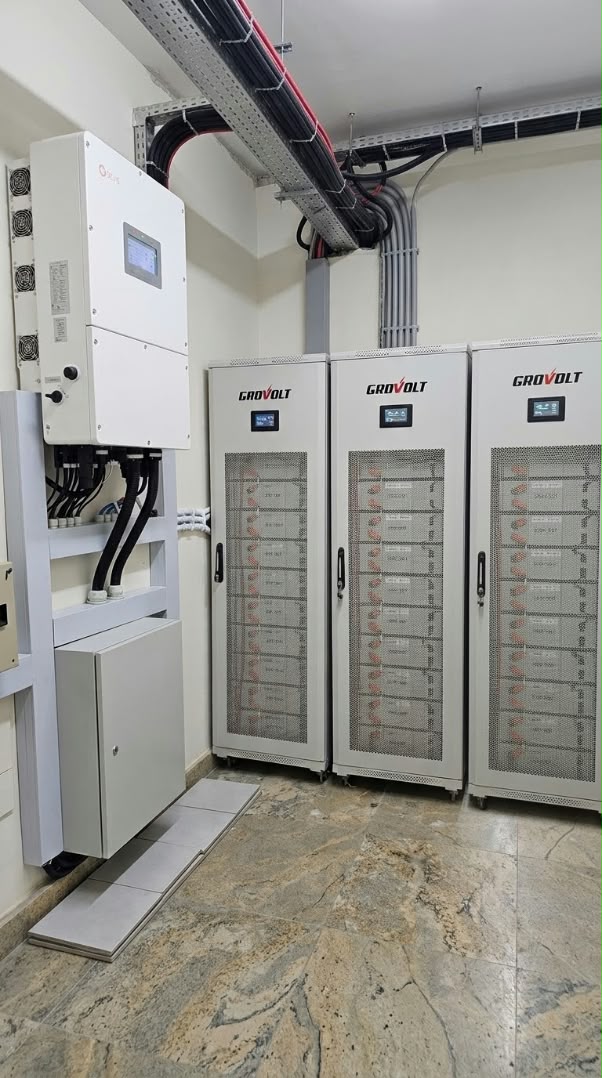 Solis three-phase hybrid inverter mounted on a wall beside three Grovolt high-voltage lithium battery cabinets, with cable trays and conduits running overhead in a tidy plant room